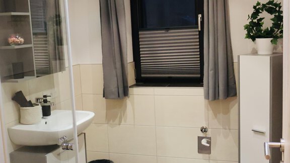 A modern bathroom with a shower, a sink, and a window. The walls are light, and the floor is equipped with dark-colored tiles. - Apart Katja - Längenfeld