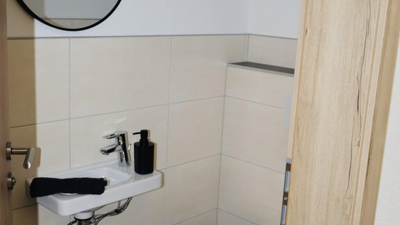 A small, modern bathroom with a sink and a toilet. The walls are tiled in light colors, and a round mirror hangs above the sink. - Apart Katja - Längenfeld