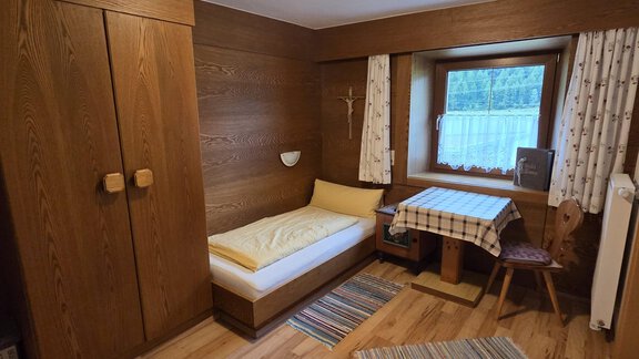 Ein gemütliches Schlafzimmer aus Holz mit einem Einzelbett und einem kleinen Tisch. Vor einem Fenster stehen Vorhänge und es gibt einen Kleiderschrank an der Wand. - Apart Luca - Längenfeld