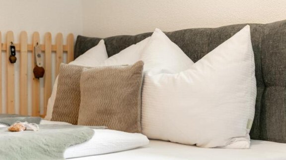 A cozy bed with several pillows in soft colors. In the background, a wooden wall is visible, giving a homely impression. - Apartment DoLa - Längenfeld