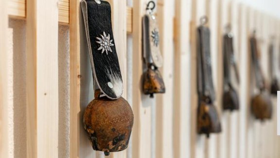 A series of traditional cowbells is hanging on a wooden slatted wall. The bells have various designs and colors. - Apartment DoLa - Längenfeld
