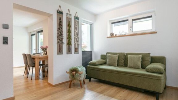 Ein helles Wohnzimmer mit einem grünen Sofa und Holzfußboden. Im Hintergrund sind ein Essbereich und dekorative Pflanzen an der Wand sichtbar. - Apartment DoLa - Längenfeld