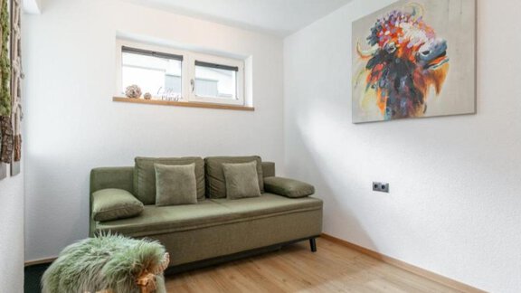 Ein modernes Wohnzimmer mit einem grünen Sofa und einem bunten Kunstwerk an der Wand. Der Raum hat helles Holzfußboden und viel natürliches Licht. - Apartment DoLa - Längenfeld
