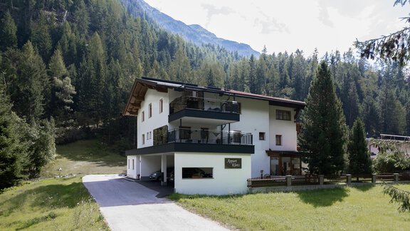 Ein modernes Gebäude in der Natur, umgeben von hohen Bäumen und Bergen. Die Umgebung ist grün und einladend. - Appart Klotz Alpenglück - Längenfeld