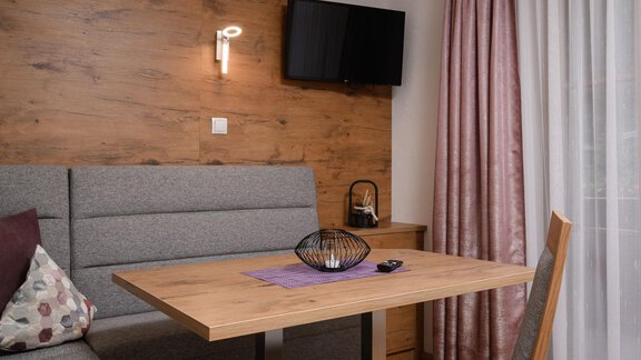 Ein modernes Wohnzimmer mit einem Holztisch und einer grauen Bank. An der Wand hängt ein Fernseher, und durch das Fenster fällt warmes Licht auf den Raum. - Appart Klotz Alpenglück - Längenfeld