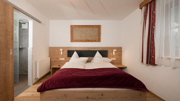 Ein gemütliches Schlafzimmer mit einem breiten Bett und zwei Kissen. Der Raum hat helle Wände und Holzdetails, sowie ein Badezimmer im Hintergrund. - Appart Klotz Alpenglück - Längenfeld