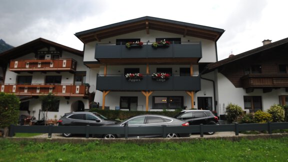 A traditional alpine building with two floors and balconies, adorned with flowers. In the foreground, some cars and a well-maintained green area can be seen. - Appart Tanja - Längenfeld