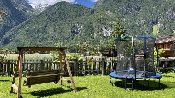 Ein schöner Garten mit einer Schaukel und einem Trampolin. Im Hintergrund erheben sich majestätische Berge unter einem blauen Himmel. - Appartement am Sonnhof - Längenfeld