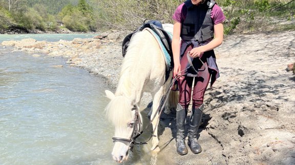 Ein Mädchen steht an einem Flussufer und führt ein weißes Pony. Es ist ein sonniger Tag mit blauem Himmel und grünen Bäumen im Hintergrund. - Appartement am Sonnhof - Längenfeld