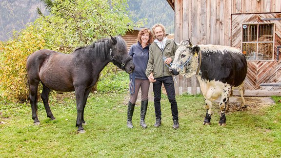 Ein Paar steht in einem Garten zwischen einem schwarzen Pferd und einer Kuh. Im Hintergrund ist eine Holzscheune und Bäume zu sehen. - Appartement am Sonnhof - Längenfeld