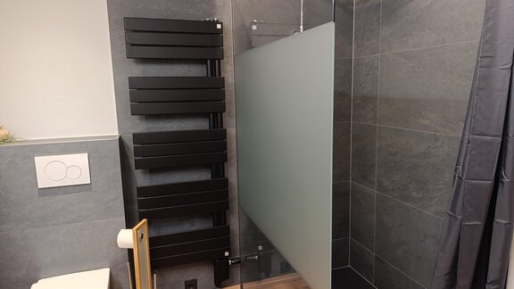 A modern bathroom with an open shower and a frosted glass partition. On the wall, there is a black towel warmer. - Appartement Thermenzeit - Längenfeld
