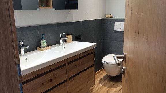 A modern bathroom with wooden cabinets and a double sink. The floor is made of wood and there is a stylish mat in front of the toilet. - Appartement Thermenzeit - Längenfeld