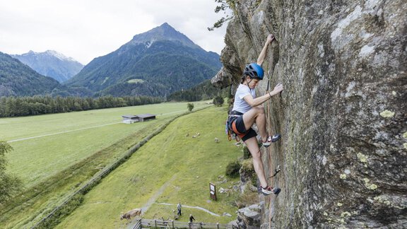 A climber in safety gear is ascending a rock face in a mountainous landscape. In the background, green meadows and mountains can be seen. - Bauernhaus Auer - Längenfeld