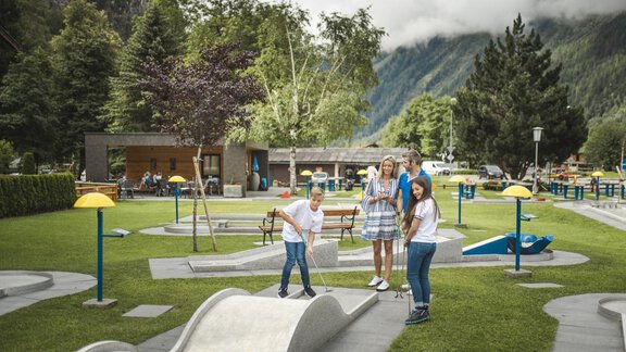 A group of four people is playing mini golf in a beautiful park. In the background, there are trees and a mountainous landscape. - Bauernhaus Auer - Längenfeld