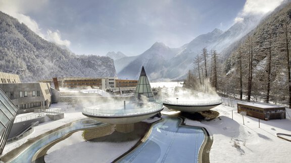 A modern thermal bath facility in a snow-covered mountain landscape. Steam clouds rise above the water basins and are surrounded by tall mountains. - Bauernhaus Auer - Längenfeld