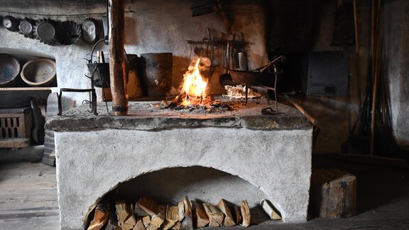 A traditional fireplace with a blazing fire in a rustic kitchen. In the background, various cooking utensils and stacks of wood are visible. - Bauernhaus Auer - Längenfeld