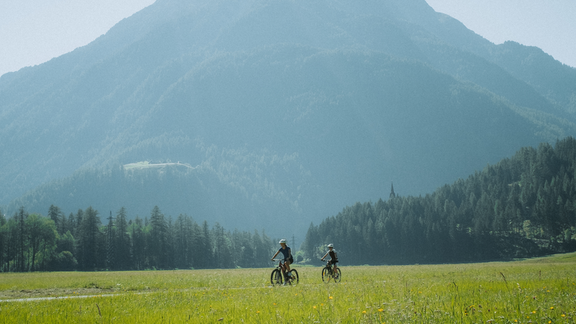 Two cyclists are riding through a green meadow. In the background, a tall mountain rises under a clear sky. - Bauernhaus Auer - Längenfeld