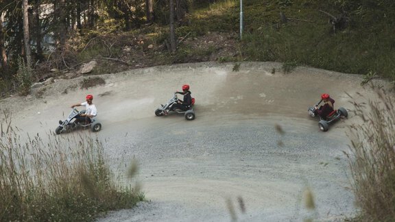 Three people are driving go-karts on a winding track in the forest. They are wearing red helmets and the surroundings are green and natural. - Bauernhaus Auer - Längenfeld