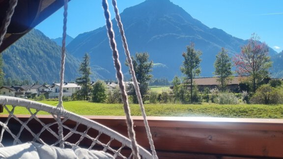 A cozy hammock with pillows, viewed from a balcony. In the background, there are tall mountains and a clear blue sky. - Bauernhaus Auer - Längenfeld