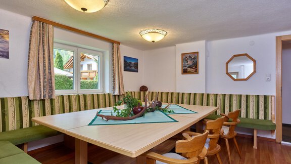 Ein gemütlicher Essbereich mit einem großen Tisch und Holzstühlen. Die Wände sind freundlich dekoriert, und große Fenster bieten Tageslicht. - Ferienhaus Schrankogelblick - Längenfeld