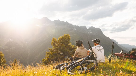 Zwei Radfahrer sitzen auf einer Wiese und genießen die Aussicht auf die Berge. Die Sonne scheint sanft und verleiht der Szene eine warme Atmosphäre. - Ferienhaus Schrankogelblick - Längenfeld