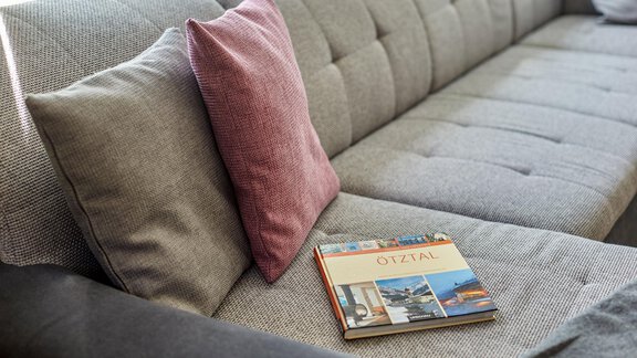 Ein modernes Sofa mit hellgrauem Stoff und einem roten Kissen. Auf dem Sofa liegt ein Buch mit dem Titel "Ötztal". - Ferienhaus Schrankogelblick - Längenfeld
