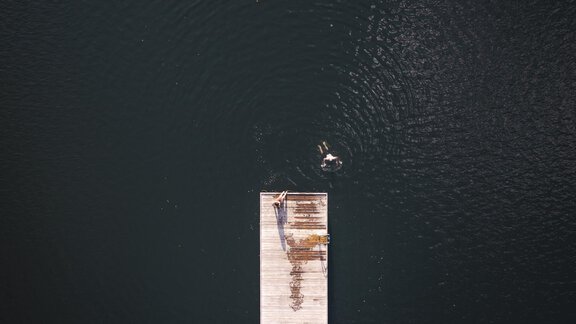 Ein Holzsteg erstreckt sich über stille, dunkle Wasseroberfläche. Eine Person schwimmt in der Nähe des Stegs. - Ferienhaus Schrankogelblick - Längenfeld