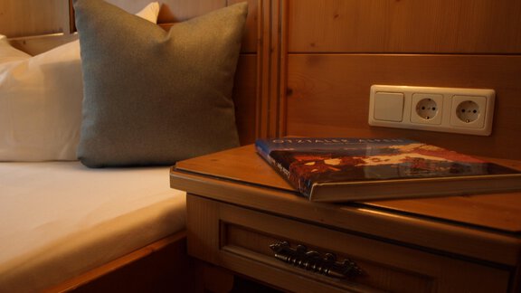 Ein gemütliches Schlafzimmer mit einem Bett und einem Kissen. Auf dem Nachttisch liegt ein Buch und es sind Steckdosen sichtbar. - Ferienhaus Schrankogelblick - Längenfeld