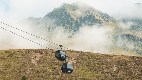 Eine Seilbahn fährt über eine grüne Berglandschaft. Im Hintergrund sind majestätische Berge und Nebel zu sehen. - Ferienhaus Schrankogelblick - Längenfeld