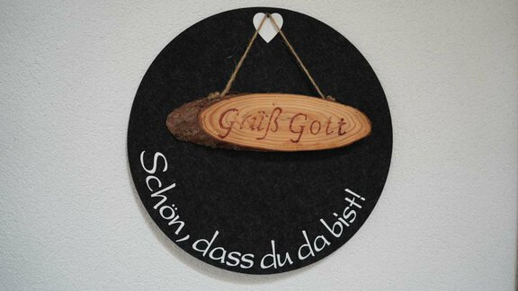 A round wall decoration in a dark color. On a wooden plaque, it says "Grüß Gott" and below it "Nice to have you here!" - Hauertalerhof - Längenfeld