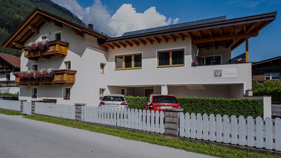 A modern house with attractive wood cladding and colorful balcony flowers. In the foreground, there are two cars and a white fence. - Haus Viktoria - Längenfeld