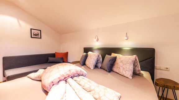 A cozy bedroom with two beds and several pillows.  
On the wall hang two lamps and there is a small seating area. - Haus Viktoria - Längenfeld