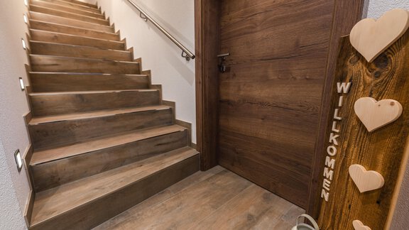 An inviting hallway with a wooden staircase and a dark front door.  
On the wall is a welcome sign with hearts. - Haus Viktoria - Längenfeld