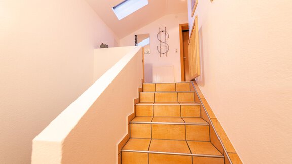 A bright staircase leads up, with brown tiles and a white railing. A window lets in light and a dollar sign hangs on the wall. - Haus Viktoria - Längenfeld