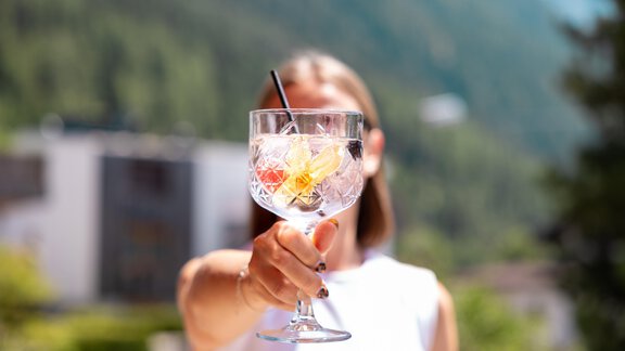 Ein erfrischender Cocktail in einem großen Glas mit Früchten und Eiswürfeln. Im Hintergrund sind Bäume und ein Gebäude zu sehen, was eine Sommeratmosphäre vermittelt. - Hotel  Bergwelt - Längenfeld