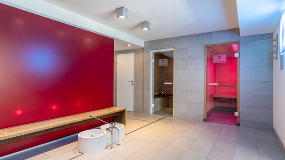 Ein moderner Wellnessbereich mit einem roten Akzentwand und Holzbank. Im Hintergrund sind zwei Saunen sichtbar, eine mit sanftem Licht. - Hotel  Bergwelt - Längenfeld