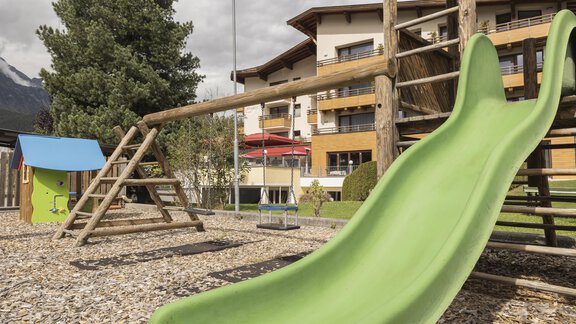 Ein Spielplatz mit einer grünen Rutsche und einer Schaukel. Im Hintergrund sind Gebäude und ein Baum zu sehen. - Hotel  Bergwelt - Längenfeld