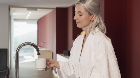 Eine entspannte Frau in einem weißen Bademantel hält eine Tasse in der modernen Küche. Der Raum hat eine helle und gemütliche Atmosphäre. - Hotel  Bergwelt - Längenfeld