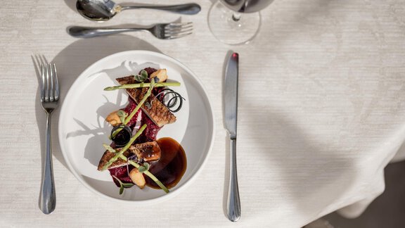 A beautifully arranged dish on a white plate. Next to it is a glass of red wine, and the cutlery is neatly placed on the table. - Hotel Rita - Längenfeld