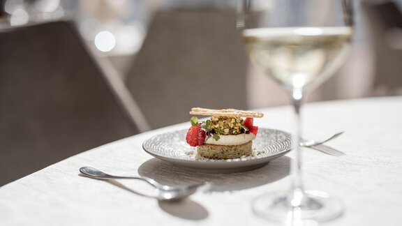 A fine dessert on a plate rim with berries and crispy topping. In the background, there is a glass of white wine. - Hotel Rita - Längenfeld