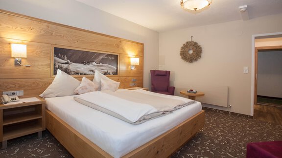 Ein modernes Hotelzimmer mit einem großen Doppelbett aus Holz und sanfter Beleuchtung. Die Einrichtung ist gemütlich mit einem Sessel und einem dekorativen Element an der Wand. - Hotel Stern - Längenfeld