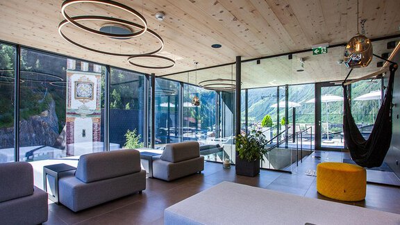 Ein modernes und helles Wohnzimmer mit großen Fenstern, die einen Blick auf die Berge bieten. Die Einrichtung besteht aus stilvollen Möbeln und hängt mit eleganten Leuchten. - Hotel Stern - Längenfeld