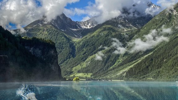 Eine Person schwimmt elegant in einem Infinity-Pool mit Blick auf majestätische Berge und einen klaren Himmel. Die Umgebung ist grün und idyllisch. - Hotel Stern - Längenfeld