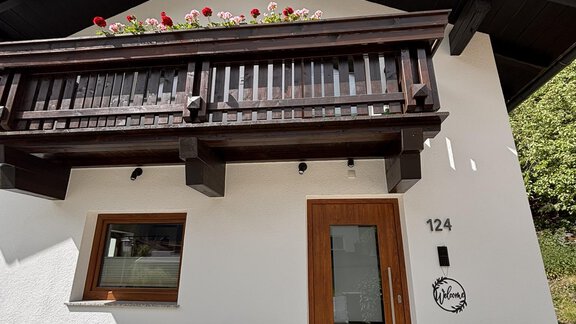 A modern, white house with a wooden balcony. The entrance area has a beautiful door and blooming flowers. - Irmas Ferienhäusl - Längenfeld