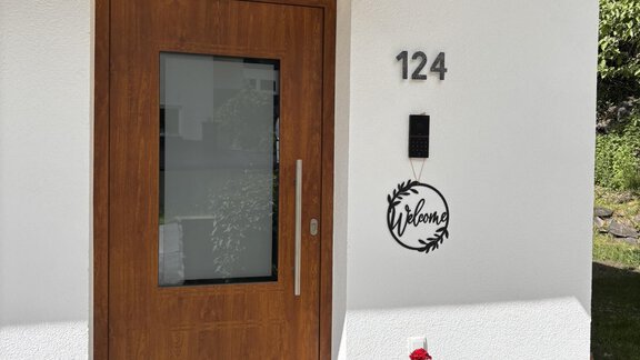 A modern house entrance with a wooden door and clean lines. Next to the door, blooming plants are in pots. - Irmas Ferienhäusl - Längenfeld