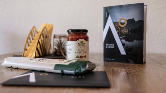 A flat surface with a glass of tomato sauce, a green package, and a brochure. In the background, decorative objects can be seen. - MYALPS Himmelreich - Längenfeld