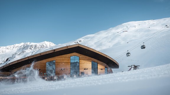 Ein modernes Gebäude in einer verschneiten Berglandschaft. Im Hintergrund sind Skilifte und schneebedeckte Berge zu sehen. - MYALPS Jordans Lodge - Längenfeld