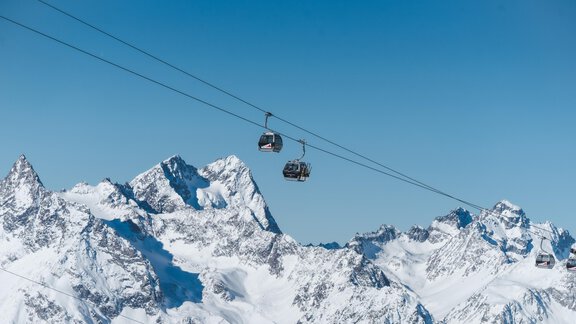 A cable car glides over snow-covered mountains beneath a clear blue sky. The majestic Alpine landscape invites skiing. - Pension Sonnenwinkl - Längenfeld