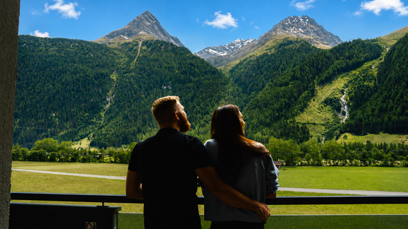 Ein Paar steht auf einem Balkon und blickt auf eine beeindruckende Berglandschaft. Hohe, grüne Berge und ein klarer Himmel schaffen eine romantische Atmosphäre. - VIER - Studios & Restaurant - Längenfeld