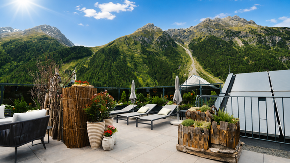 Eine Terrasse mit Liegen und Pflanzen, umgeben von majestätischen Bergen. Der Himmel ist klar und die Sonne scheint hell. - VIER - Studios & Restaurant - Längenfeld